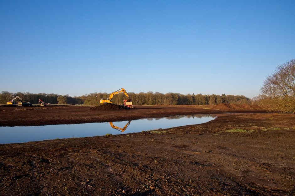 A construction site outdoors near Richmond Park showing a large muddy area with dark, moist soil and patches of water reflecting the clear blue sky. In the background, several yellow excavators and co